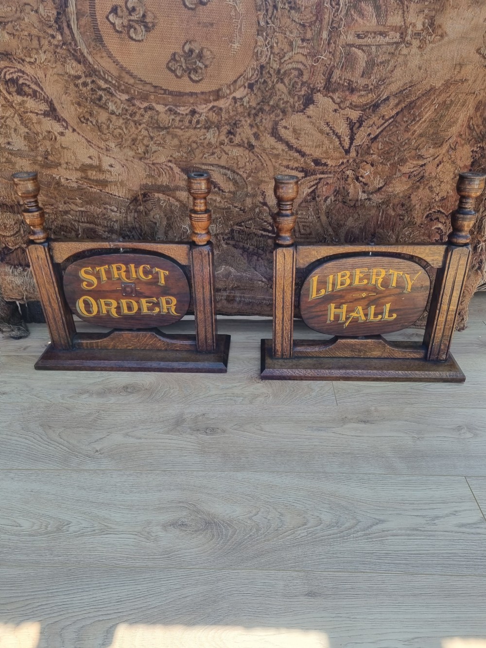 A pair of carved dark oak tabletop signs with oval central plaques featuring painted gold lettering reading Liberty Hall and Strict Order. View II