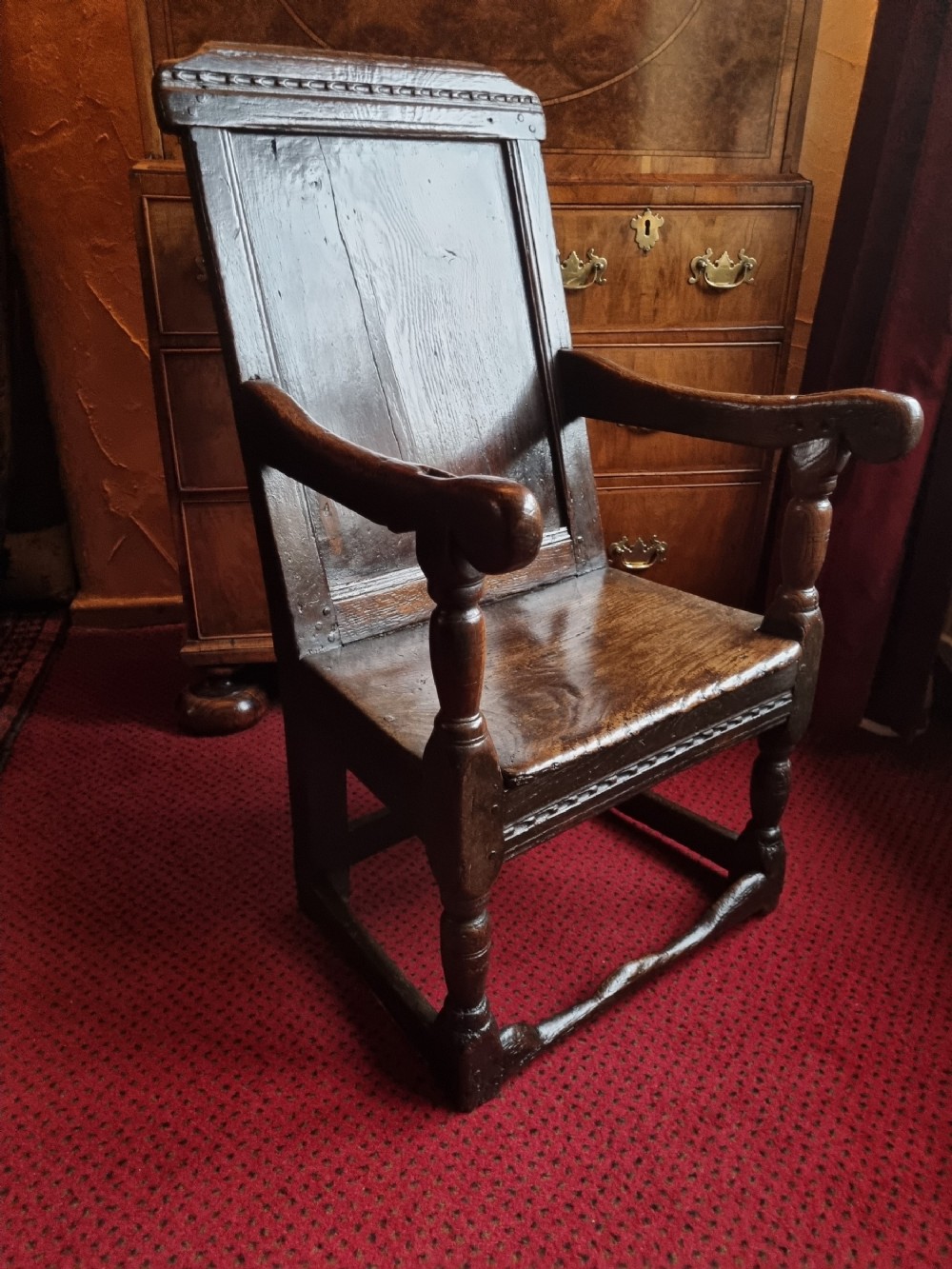 A seventeenth-century dark oak wainscot armchair featuring a solid panelled backrest, carved top rail, curved armrests, and turned leg supports.