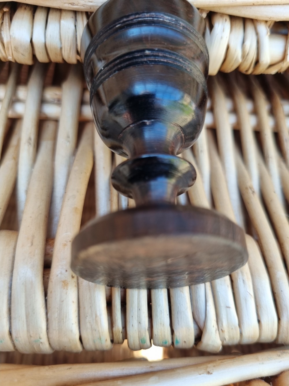 A small turned solid ebony goblet featuring a rounded bowl and a stepped foot base, set against a wicker background. View V