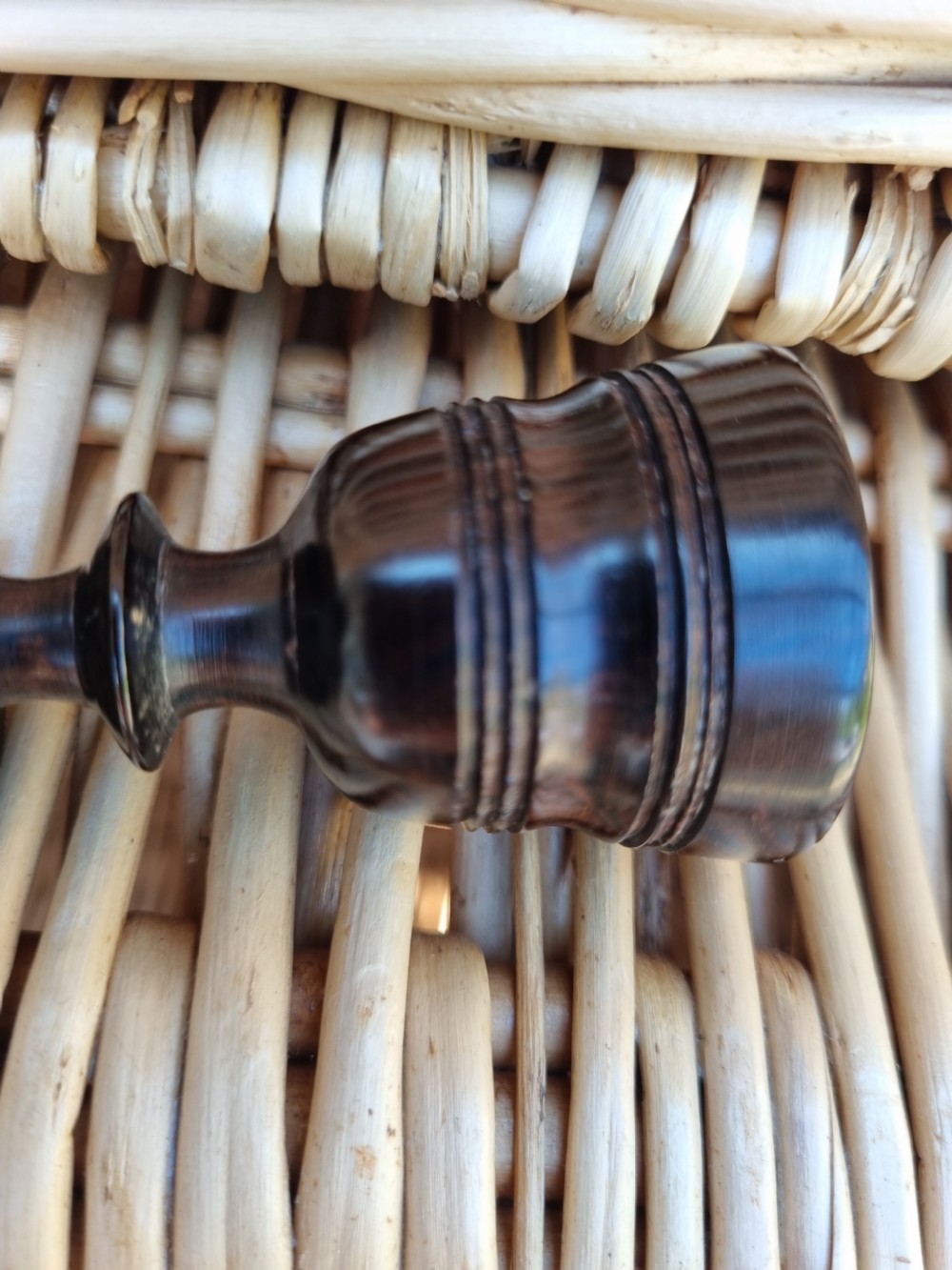 A small turned solid ebony goblet featuring a rounded bowl and a stepped foot base, set against a wicker background. View VI