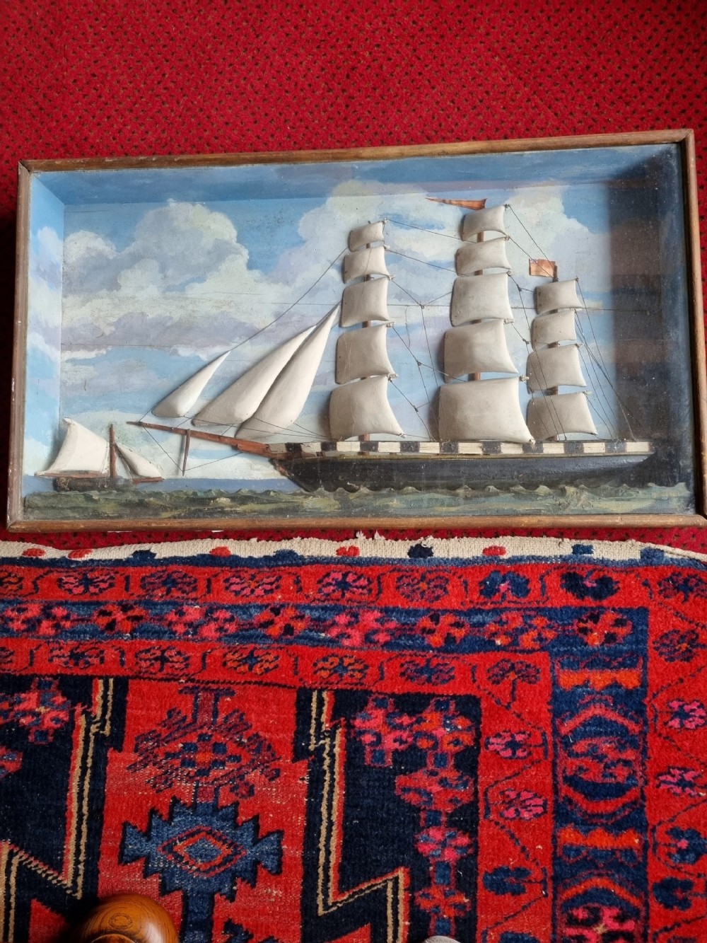 A framed three-dimensional naive wooden diorama depicting a multi-masted sailing ship on a painted sea against a cloudy sky background.
