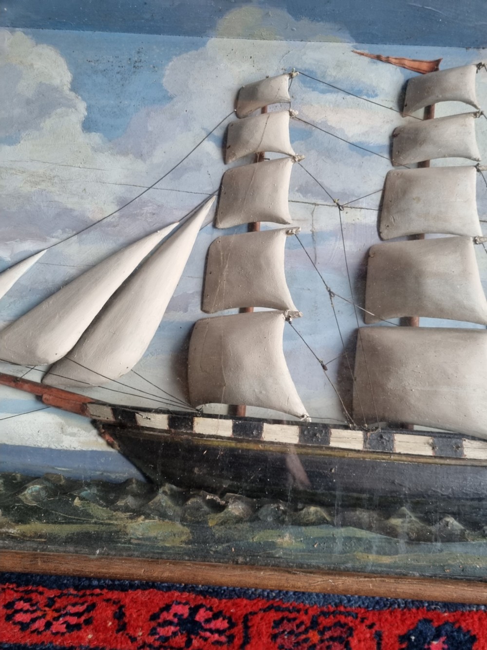 A framed three-dimensional naive wooden diorama depicting a multi-masted sailing ship on a painted sea against a cloudy sky background. View III