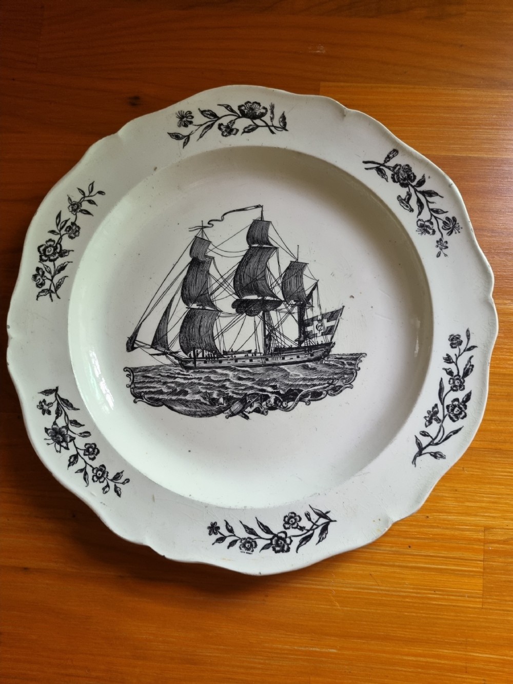 A late eighteenth or early nineteenth-century scalloped creamware plate featuring a central black transfer-printed sailing ship surrounded by floral border sprays.