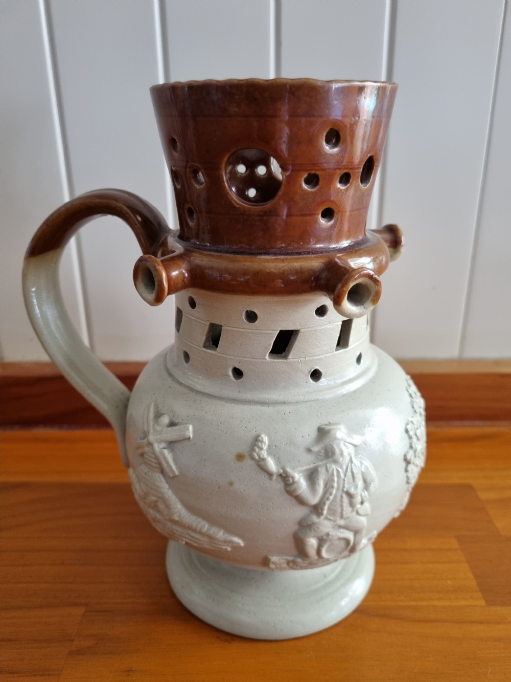A 19th-century salt-glazed stoneware puzzle jug featuring a brown pierced upper neck, a cream body with a windmill relief decoration, and a hollow loop handle. View VII