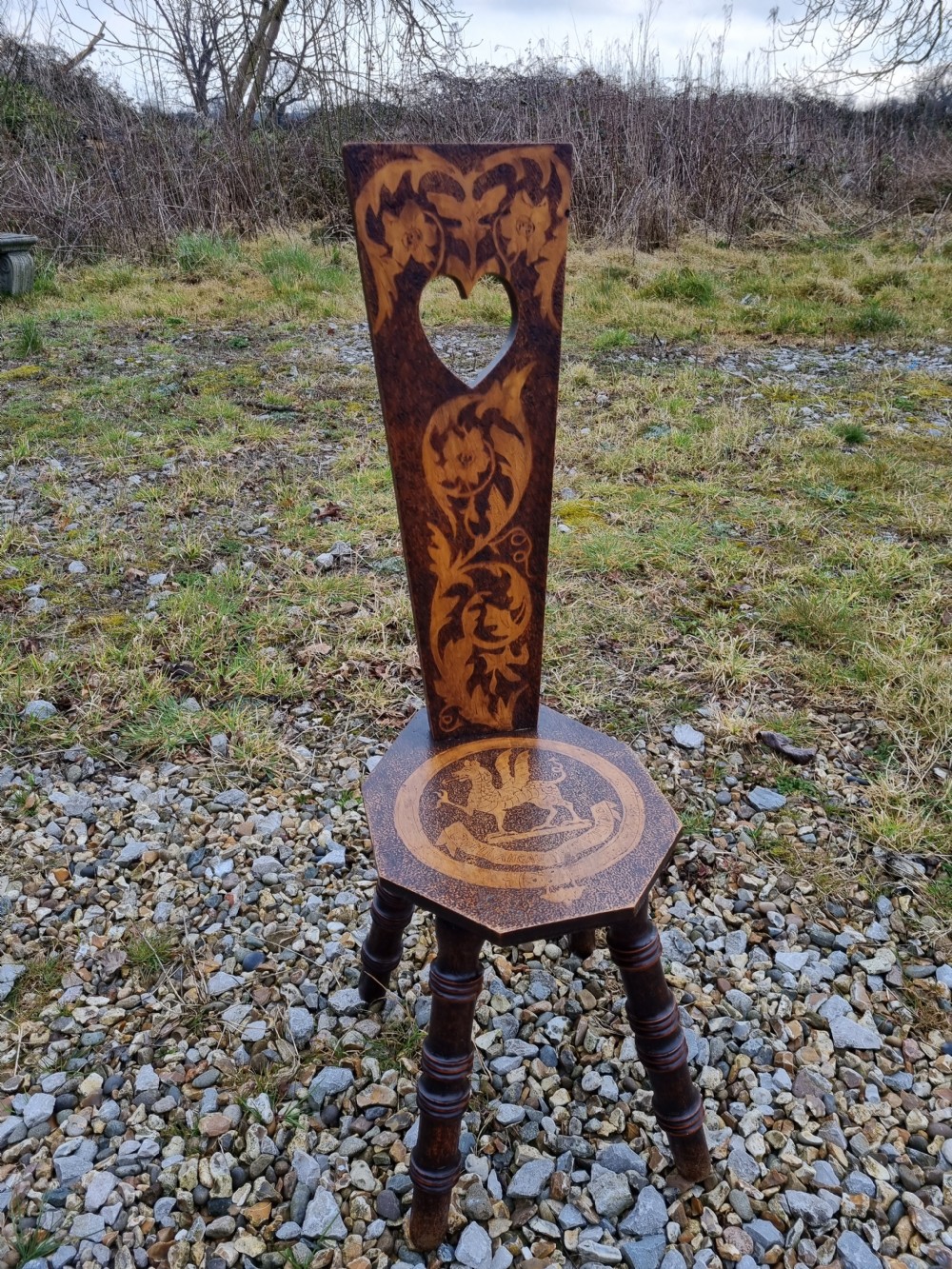 A 19th-century Welsh folk art spinning chair with a tall back featuring a heart cut-out, floral pyrography, a hexagonal seat with a carved dragon motif, and four turned legs. View VI