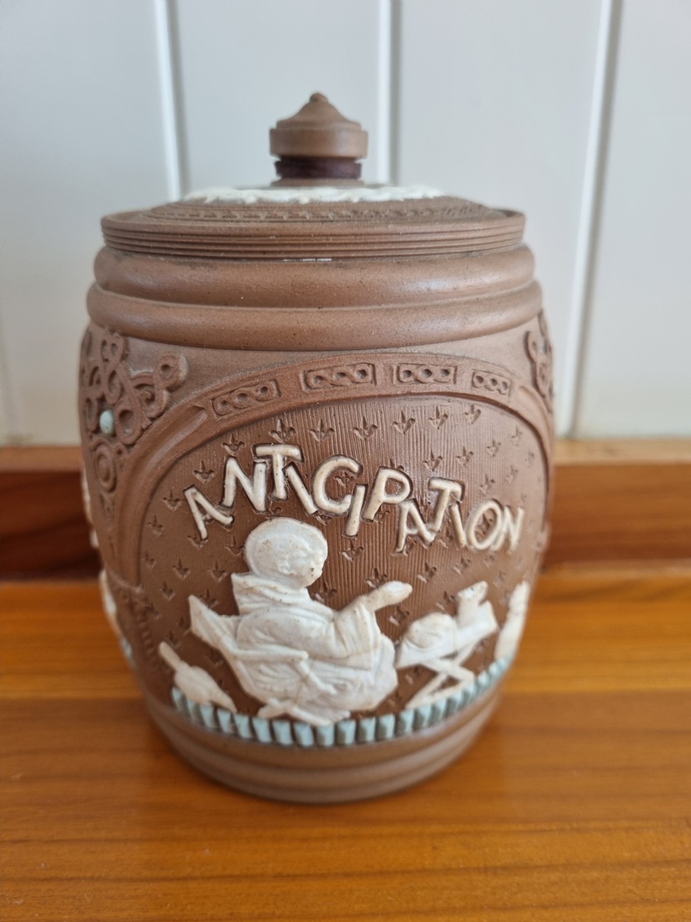 A cylindrical brown stoneware biscuit jar with a finial lid, featuring white sprigged relief lettering reading Anticipation above a figural scene.