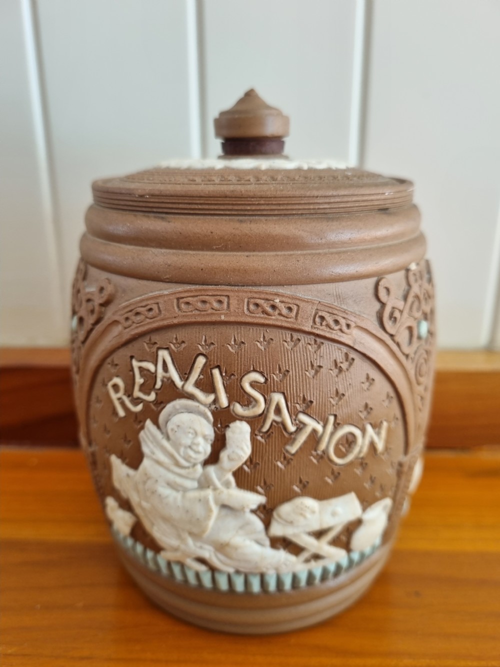 A cylindrical brown stoneware biscuit jar with a finial lid, featuring white sprigged relief lettering reading Anticipation above a figural scene. View II