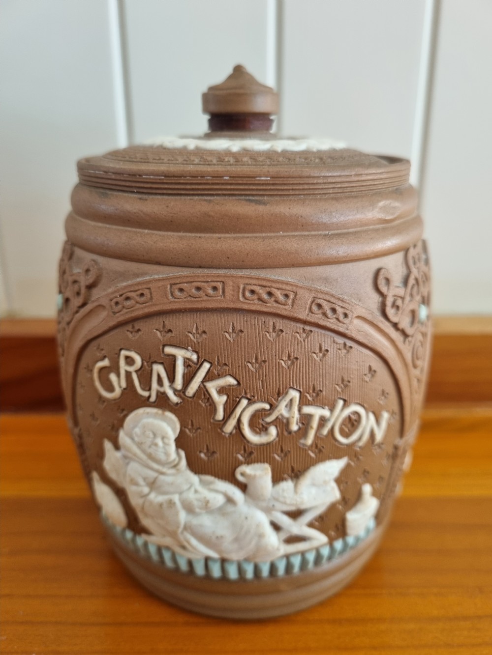 A cylindrical brown stoneware biscuit jar with a finial lid, featuring white sprigged relief lettering reading Anticipation above a figural scene. View III