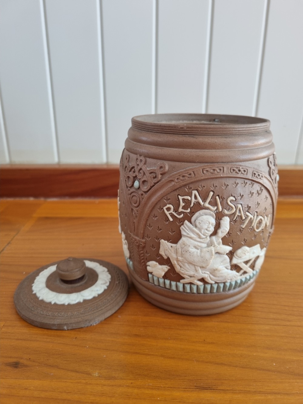 A cylindrical brown stoneware biscuit jar with a finial lid, featuring white sprigged relief lettering reading Anticipation above a figural scene. View VI