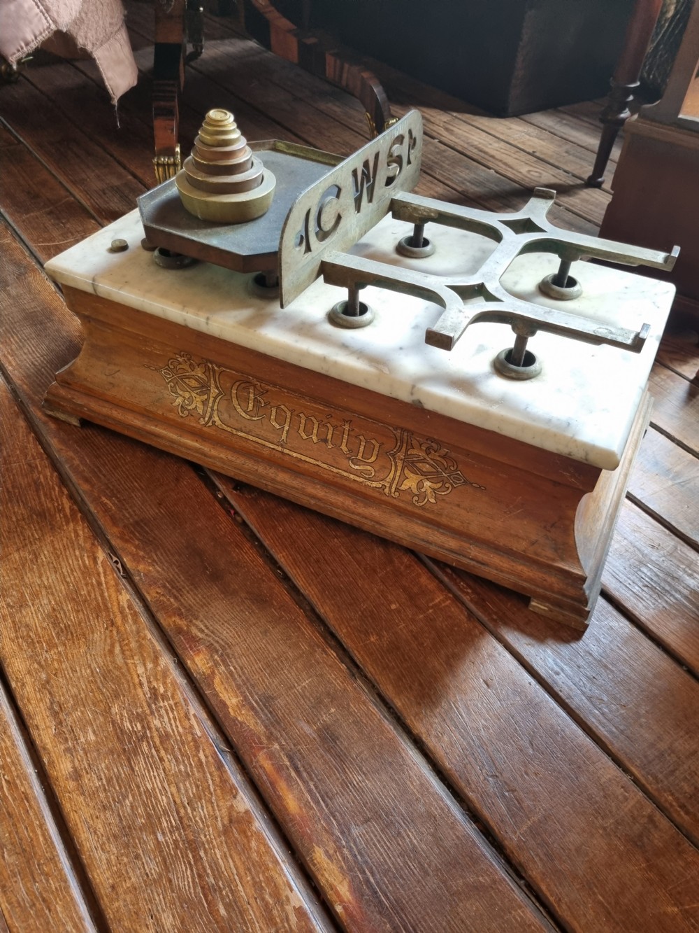 A Victorian grocer balance scale featuring a wooden base with gilded Equity lettering, a white marble slab surface, cast metal framework marked CWS, and nested brass weights. View VII