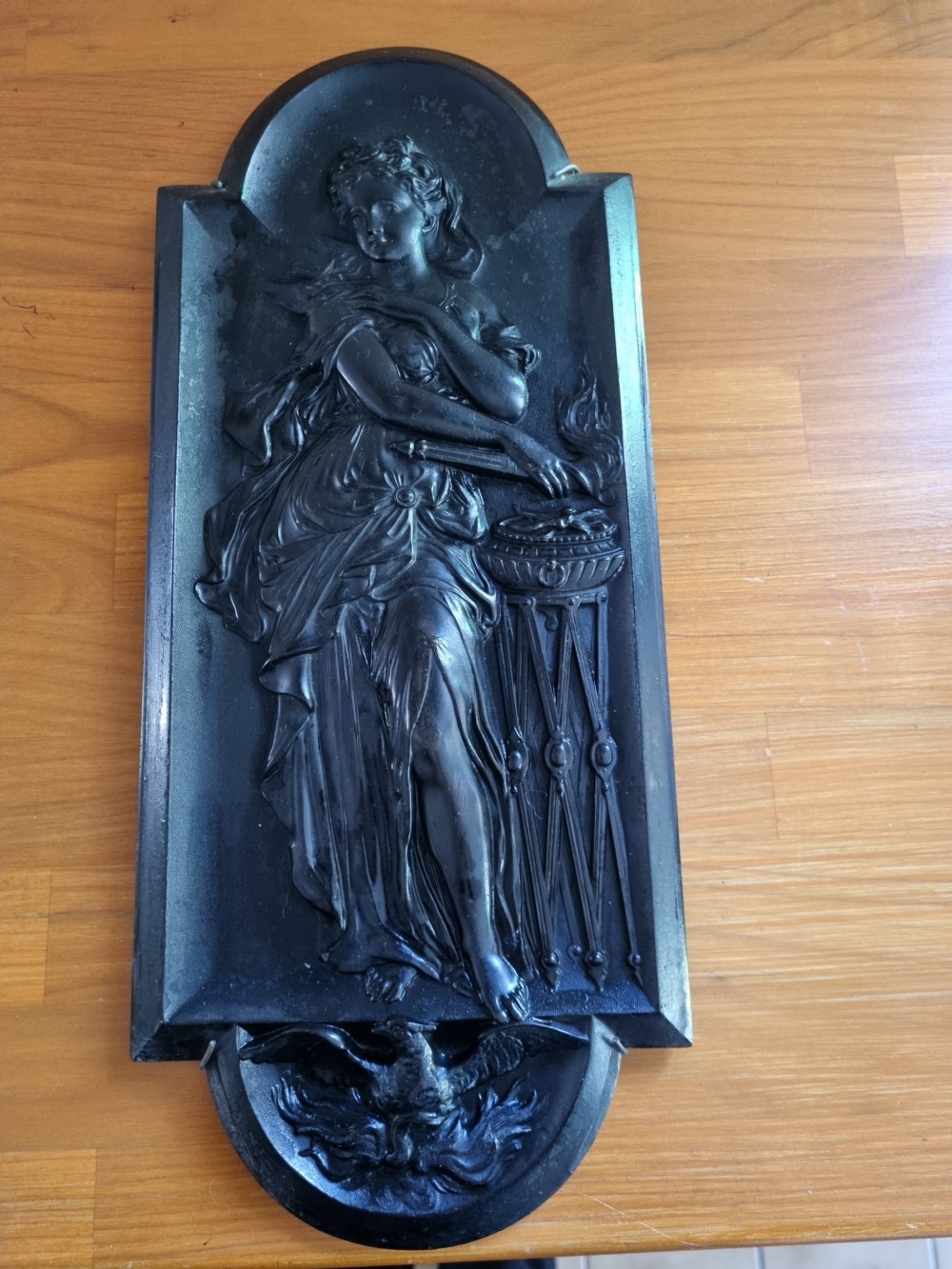A dark ebonised vertical relief wall plaque depicting the Roman goddess Vesta with classical drapery standing beside a tripod burner with an eagle motif at the base. View VII