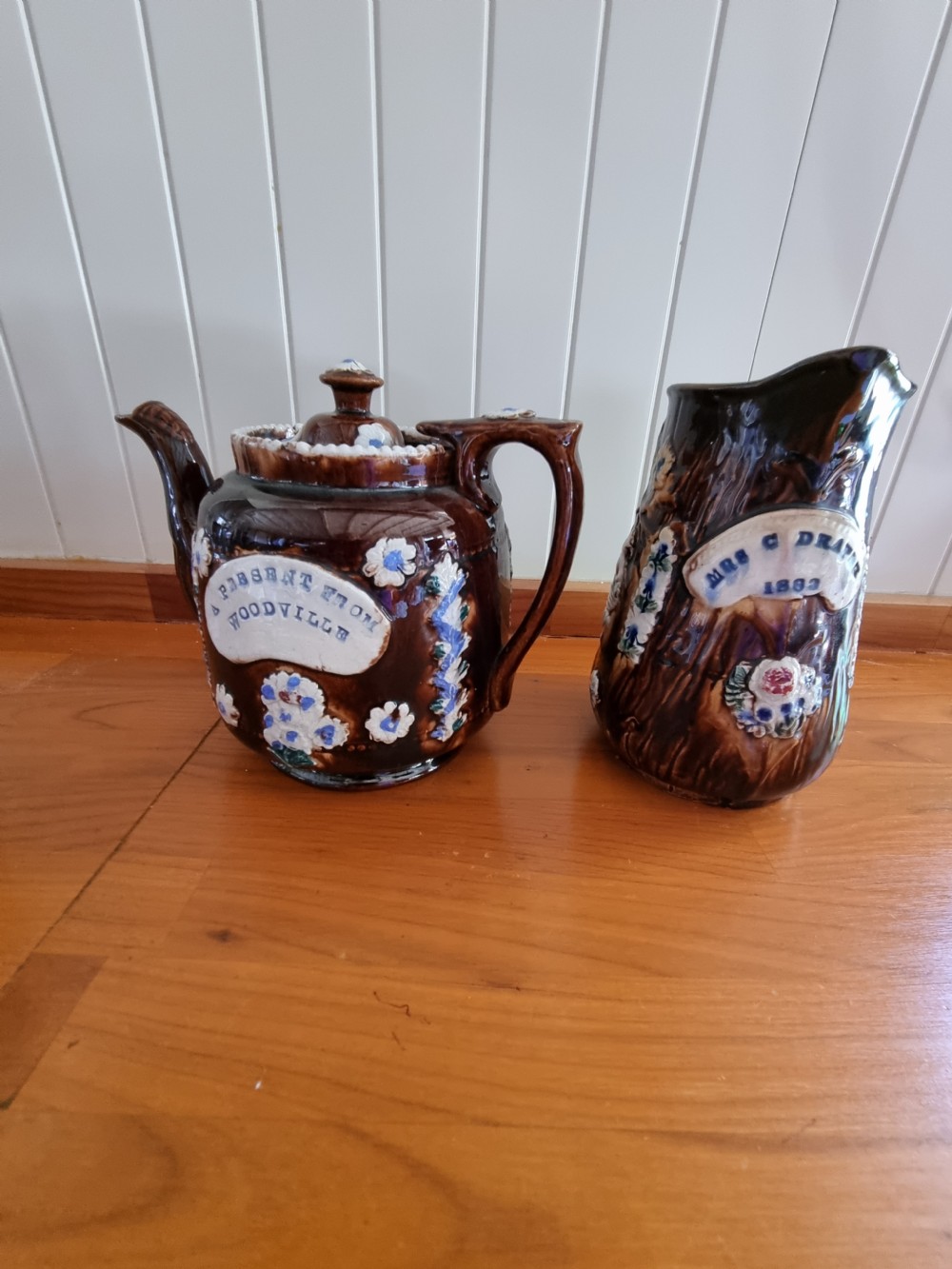 A pair of Victorian Measham Bargeware stoneware vessels, including a teapot inscribed A Present From Woodville and a jug inscribed Mrs C Drake 1893, both featuring dark brown treacle glaze and sprigged floral decoration. View V