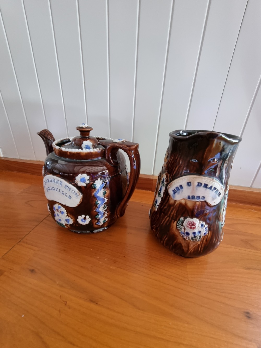 A pair of Victorian Measham Bargeware stoneware vessels, including a teapot inscribed A Present From Woodville and a jug inscribed Mrs C Drake 1893, both featuring dark brown treacle glaze and sprigged floral decoration. View VII