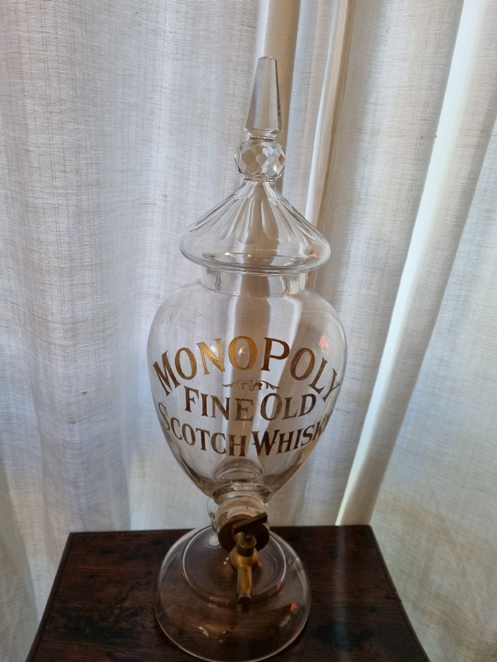 A clear glass Victorian whisky dispenser featuring gilded lettering reading Monopoly Fine Old Scotch Whisky, a brass spigot tap, and a faceted glass spire stopper.