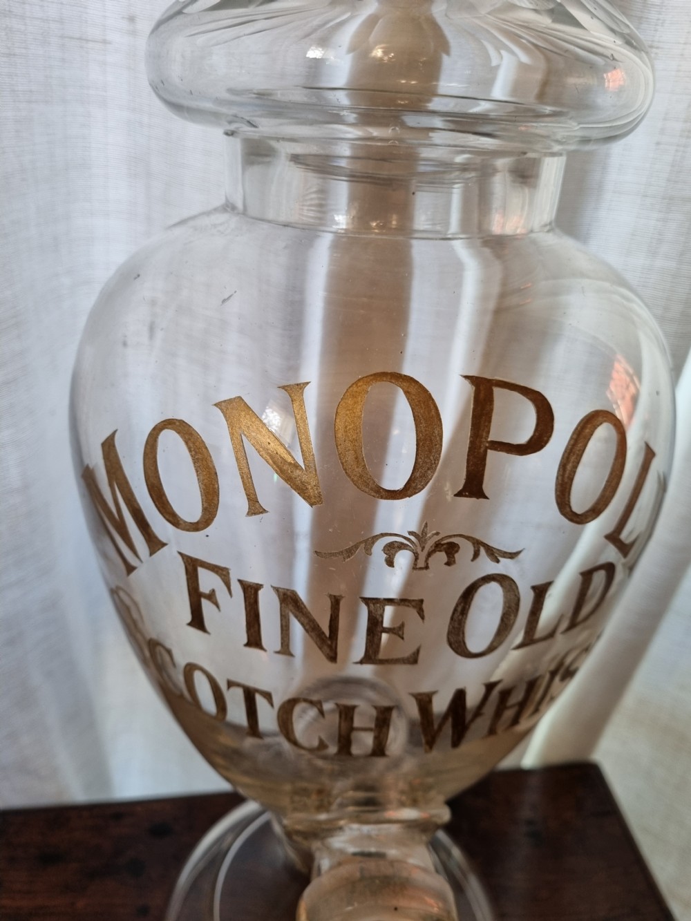 A clear glass Victorian whisky dispenser featuring gilded lettering reading Monopoly Fine Old Scotch Whisky, a brass spigot tap, and a faceted glass spire stopper. View II