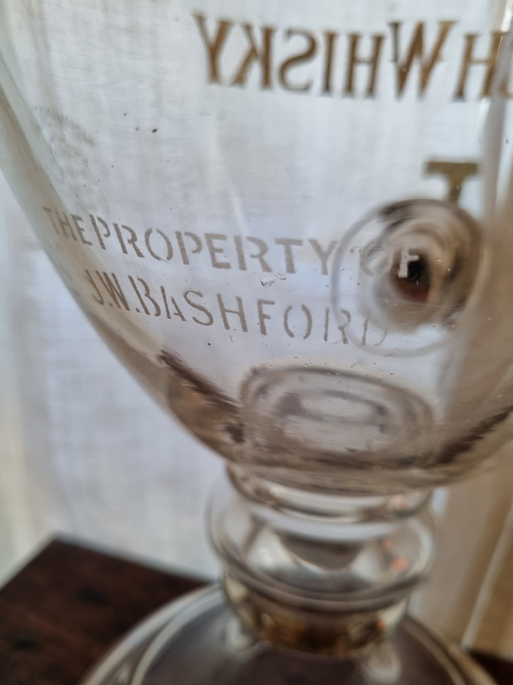 A clear glass Victorian whisky dispenser featuring gilded lettering reading Monopoly Fine Old Scotch Whisky, a brass spigot tap, and a faceted glass spire stopper. View IV