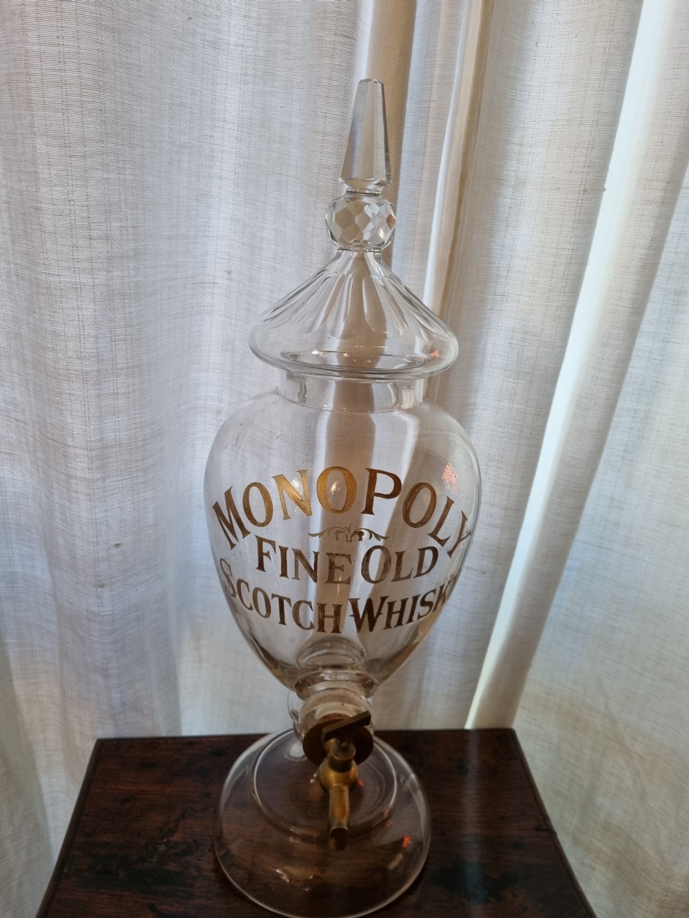 A clear glass Victorian whisky dispenser featuring gilded lettering reading Monopoly Fine Old Scotch Whisky, a brass spigot tap, and a faceted glass spire stopper. View VI