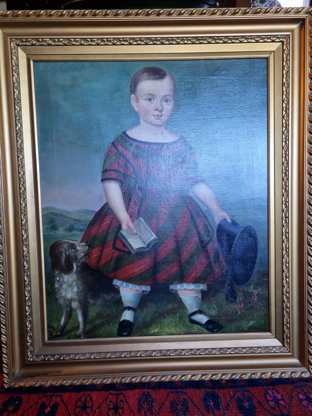 A 19th-century naive oil painting on canvas depicting a standing child in a red and dark blue striped dress with a dog, displayed within a gilded frame. View VII