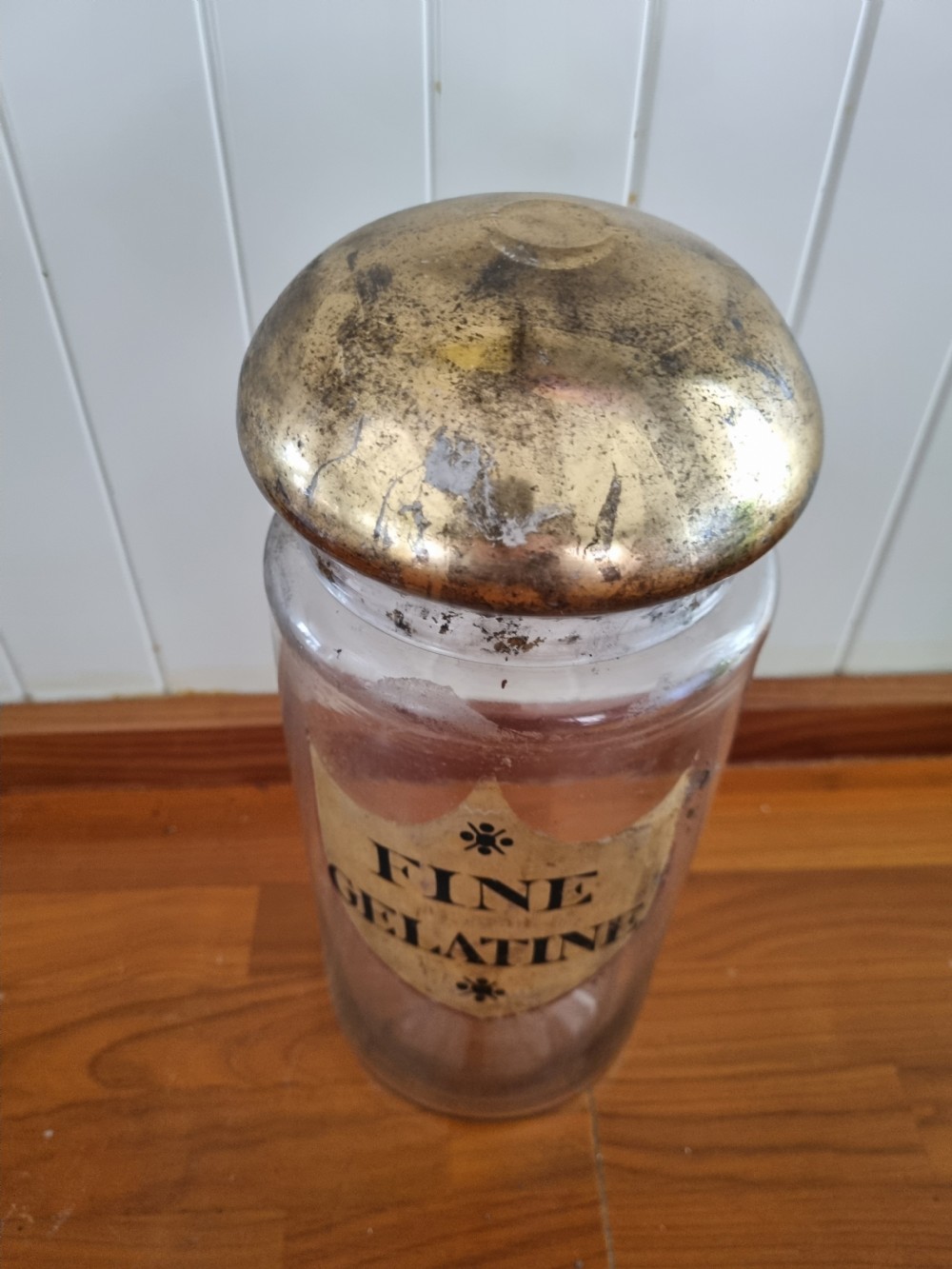 A tall clear glass chemist jar with a domed gold-toned cap and a gold shield label painted with the words Fine Gelatine. View IV