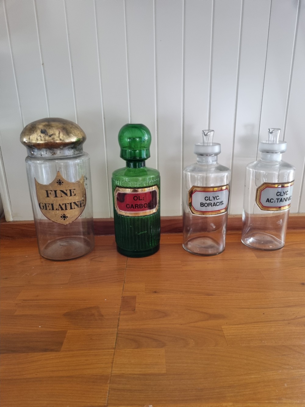 A tall clear glass chemist jar with a domed gold-toned cap and a gold shield label painted with the words Fine Gelatine. View V