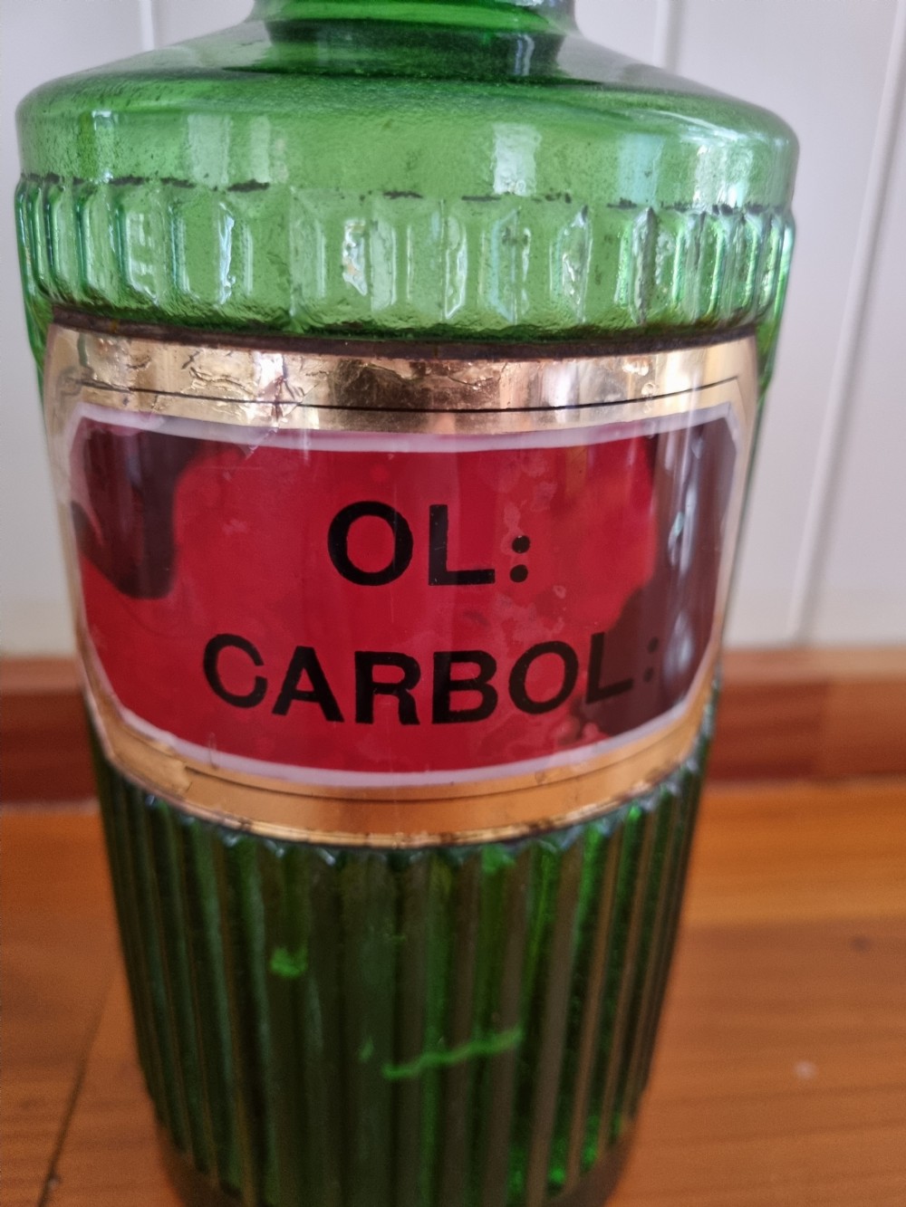 A tall 19th-century emerald green glass chemist jar featuring a ground glass stopper, fluted base, and a red gilded shield label. View IV