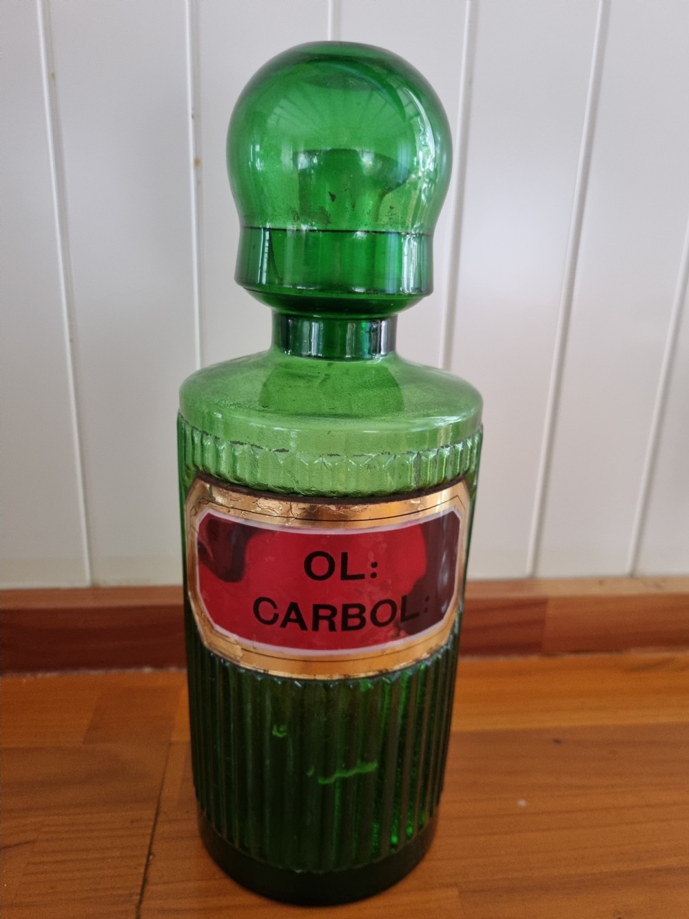 A tall 19th-century emerald green glass chemist jar featuring a ground glass stopper, fluted base, and a red gilded shield label.