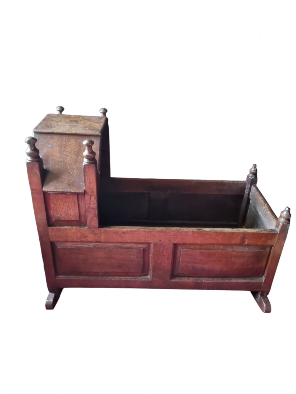 A seventeenth-century dark oak hooded cradle featuring panelled side construction, turned finials on the corner posts, and solid wooden rockers. View III