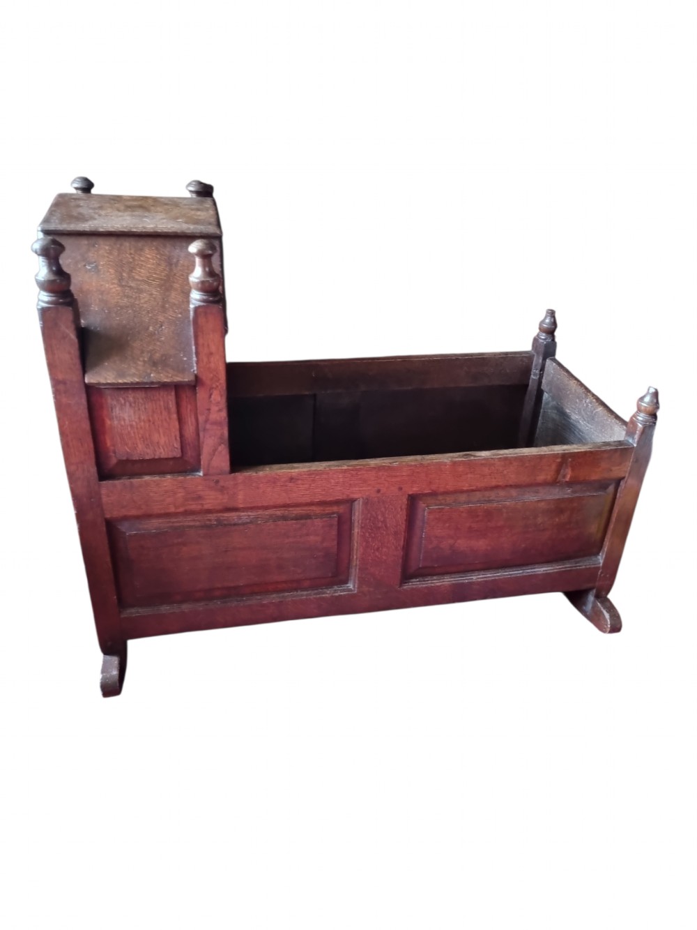 A seventeenth-century dark oak hooded cradle featuring panelled side construction, turned finials on the corner posts, and solid wooden rockers. View II