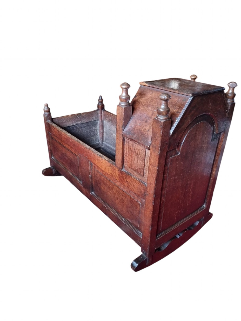 A seventeenth-century dark oak hooded cradle featuring panelled side construction, turned finials on the corner posts, and solid wooden rockers. View VII