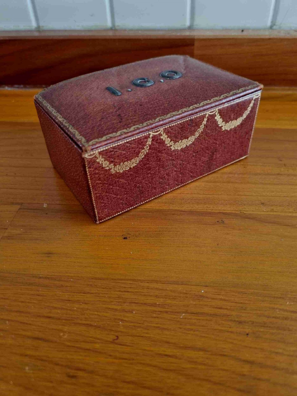 A small rectangular deep red Morocco leather stamp box with gilt festoon decoration and applied metal initials on the lid.