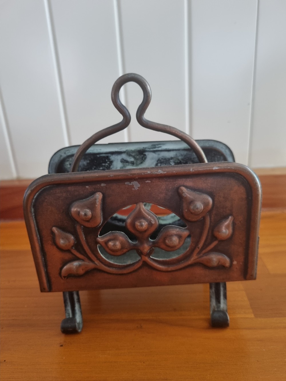 A copper Arts and Crafts letter rack featuring embossed floral motifs, a looped top handle, and four curved legs on a wooden surface. View II