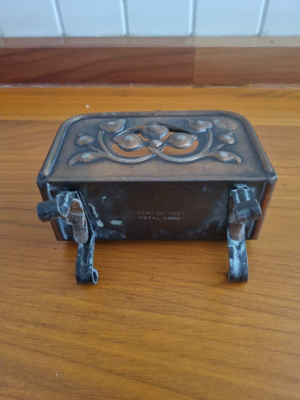 A copper Arts and Crafts letter rack featuring embossed floral motifs, a looped top handle, and four curved legs on a wooden surface. View VI