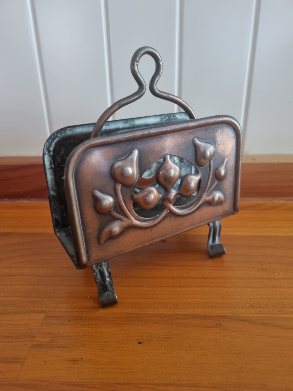 A copper Arts and Crafts letter rack featuring embossed floral motifs, a looped top handle, and four curved legs on a wooden surface. View VII