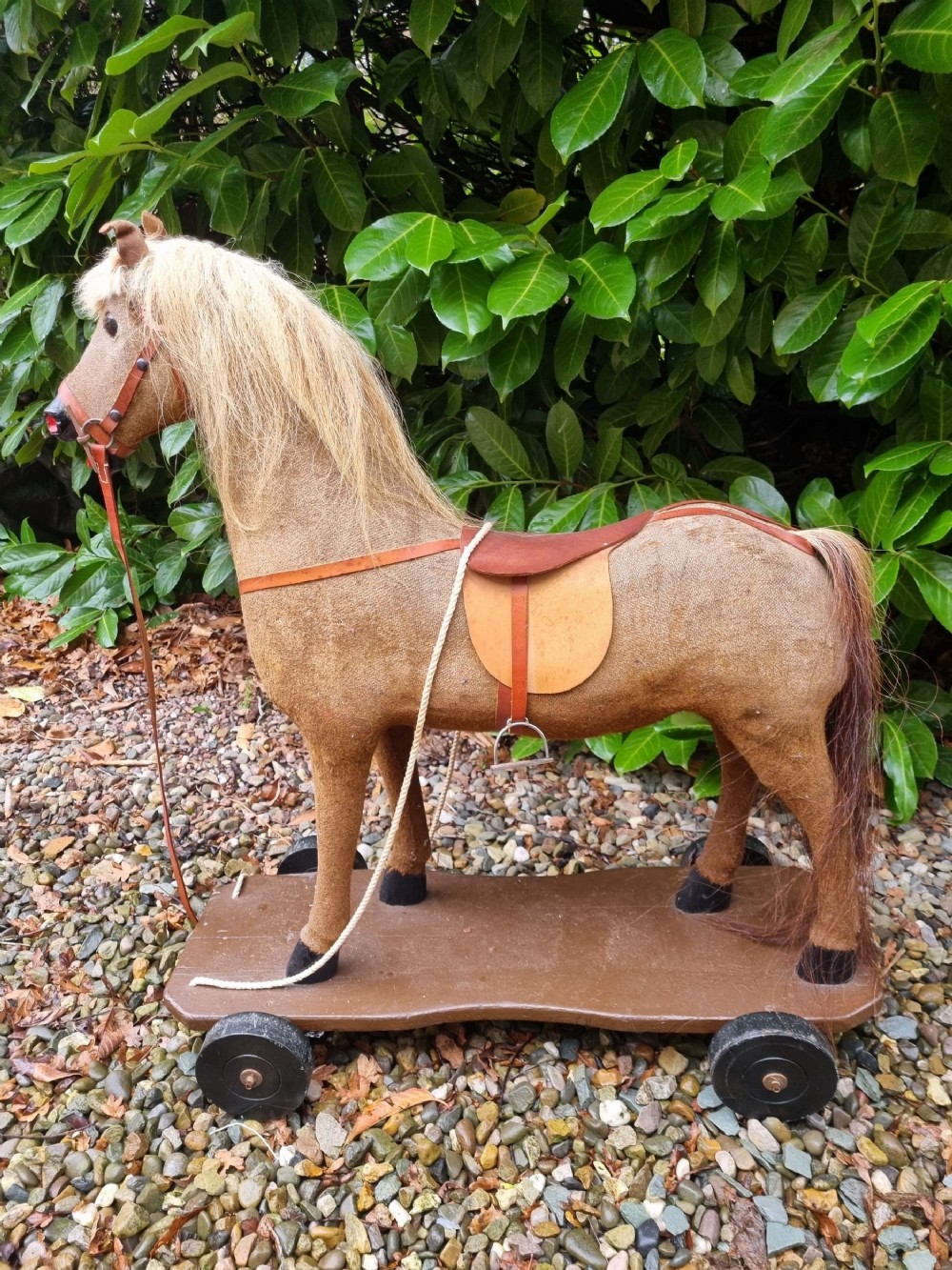 A late 19th-century childs pull-along toy horse with a tan fabric body, blonde mane, leather saddle, and bridle mounted on a brown wooden plinth with four wheels.