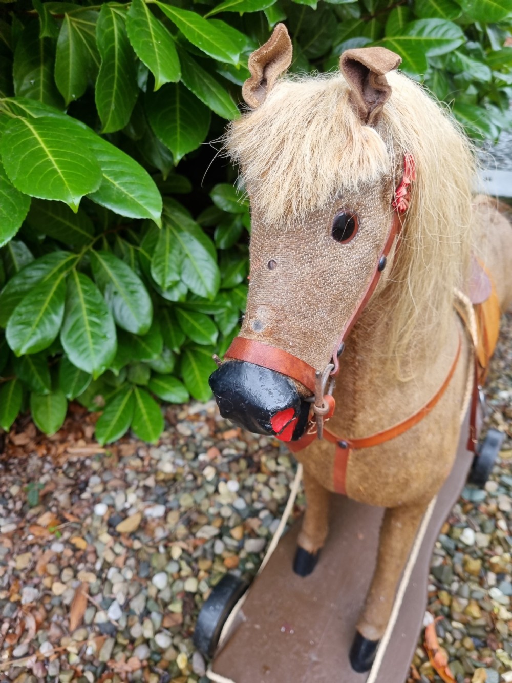 A late 19th-century childs pull-along toy horse with a tan fabric body, blonde mane, leather saddle, and bridle mounted on a brown wooden plinth with four wheels. View V