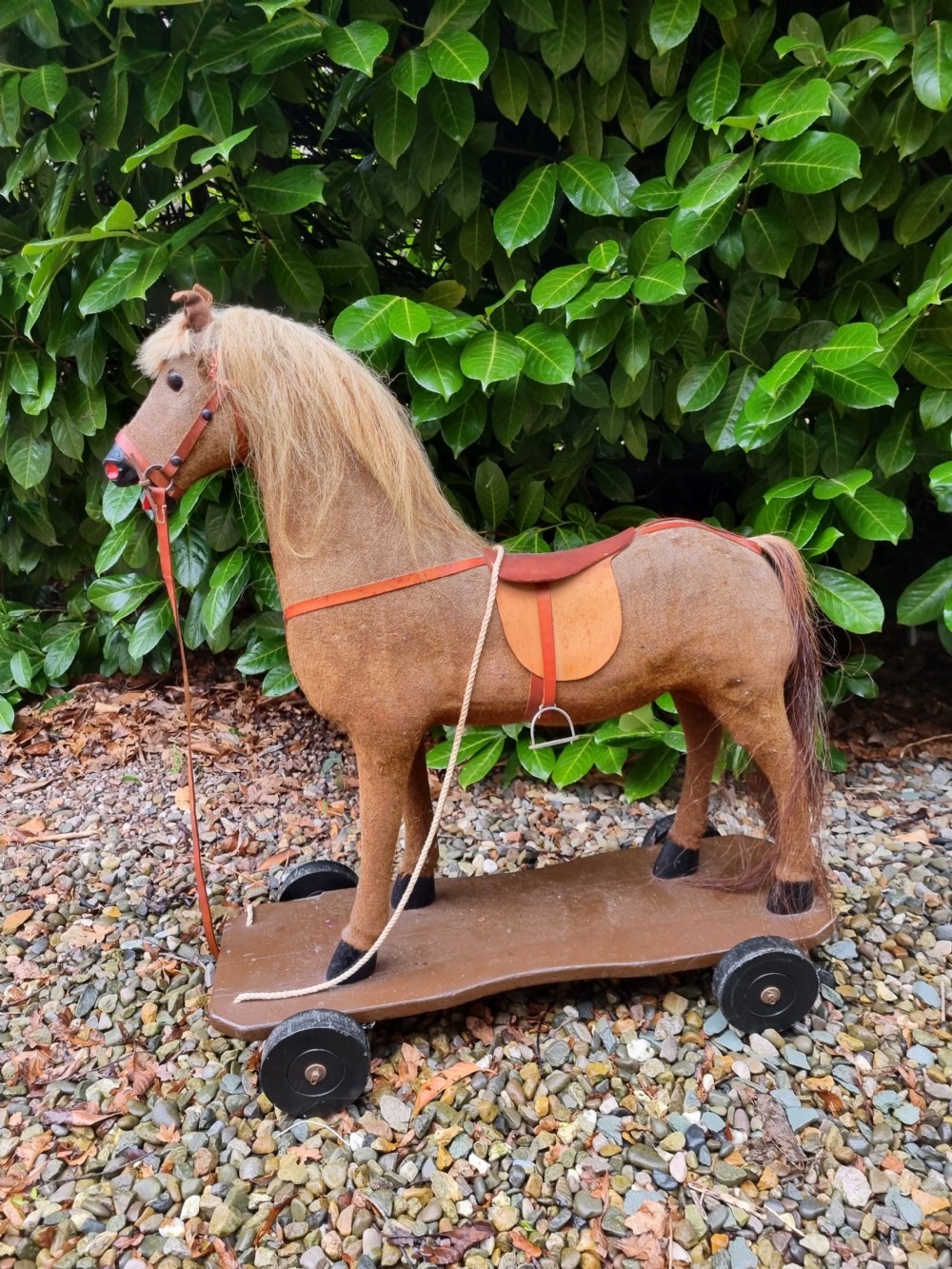 A late 19th-century childs pull-along toy horse with a tan fabric body, blonde mane, leather saddle, and bridle mounted on a brown wooden plinth with four wheels. View VIII
