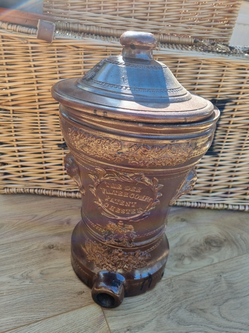A dark brown glazed stoneware water filter featuring decorative relief moulding, a domed lid, maker markings reading Dee Filter Company Chester, and a spigot opening at the base. View V