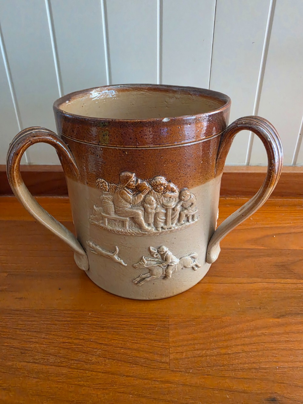 An early nineteenth-century two-tone salt-glazed stoneware tyg featuring three ribbed loop handles and raised relief sprigged decorations of tavern scenes and hunting hounds.