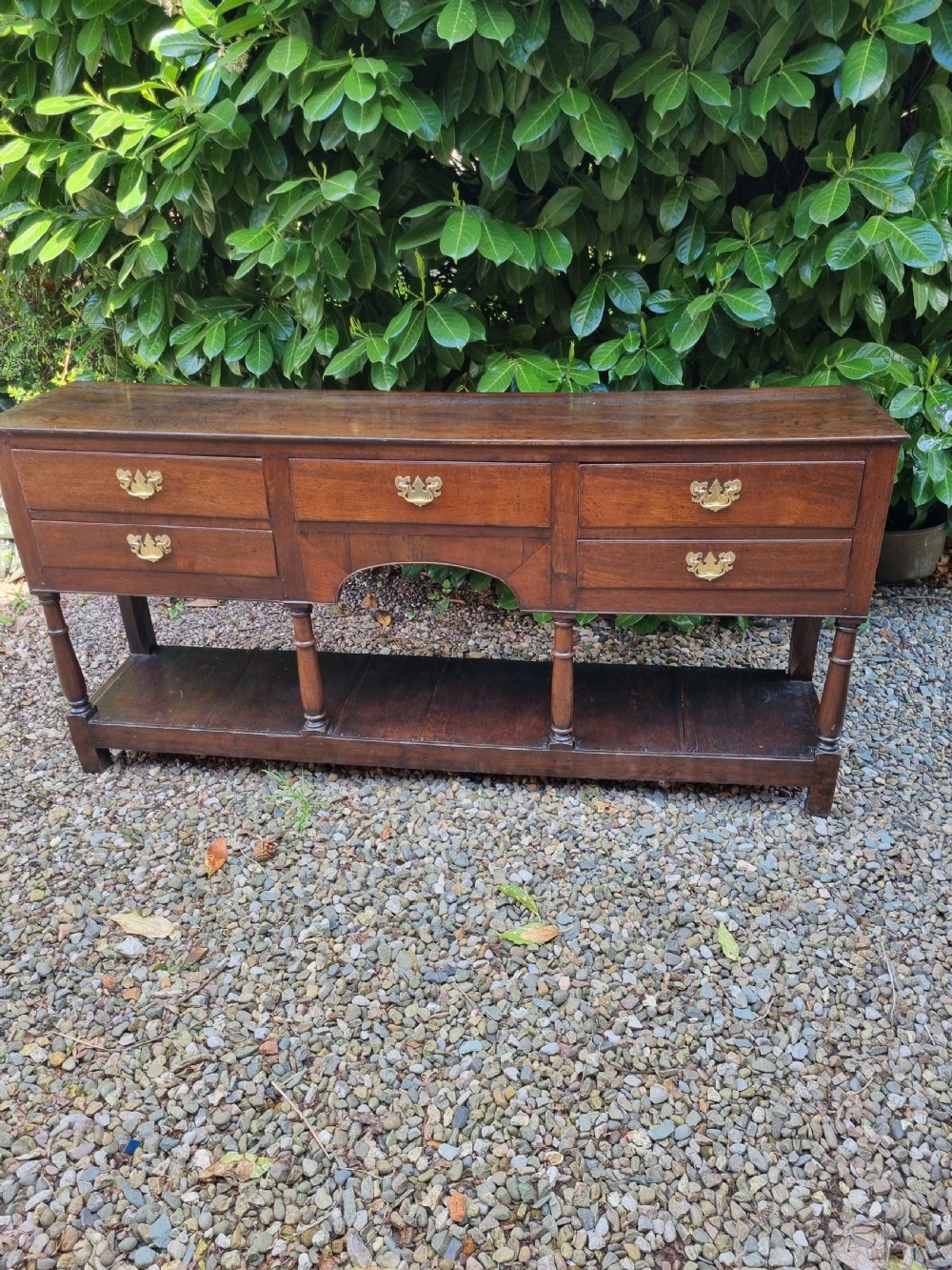 An eighteenth-century dark oak Welsh dresser base featuring five drawers with brass swan-neck handles, an arched central apron, turned supports, and a lower pot board shelf.