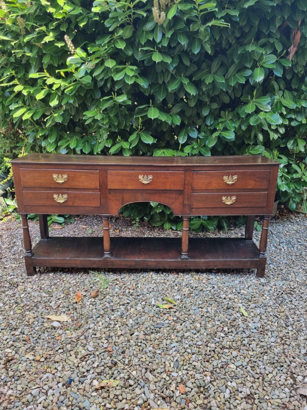An eighteenth-century dark oak Welsh dresser base featuring five drawers with brass swan-neck handles, an arched central apron, turned supports, and a lower pot board shelf. View II
