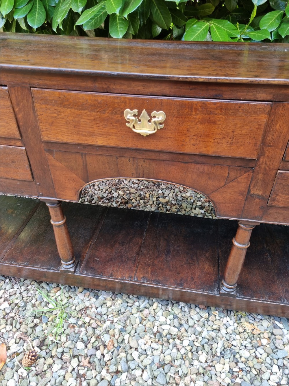 An eighteenth-century dark oak Welsh dresser base featuring five drawers with brass swan-neck handles, an arched central apron, turned supports, and a lower pot board shelf. View III