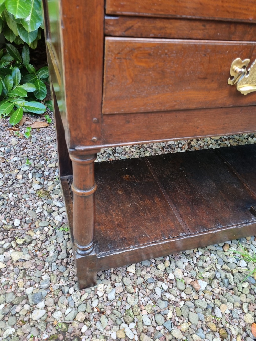 An eighteenth-century dark oak Welsh dresser base featuring five drawers with brass swan-neck handles, an arched central apron, turned supports, and a lower pot board shelf. View IV