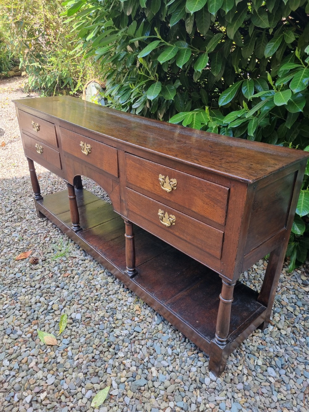 An eighteenth-century dark oak Welsh dresser base featuring five drawers with brass swan-neck handles, an arched central apron, turned supports, and a lower pot board shelf. View VII