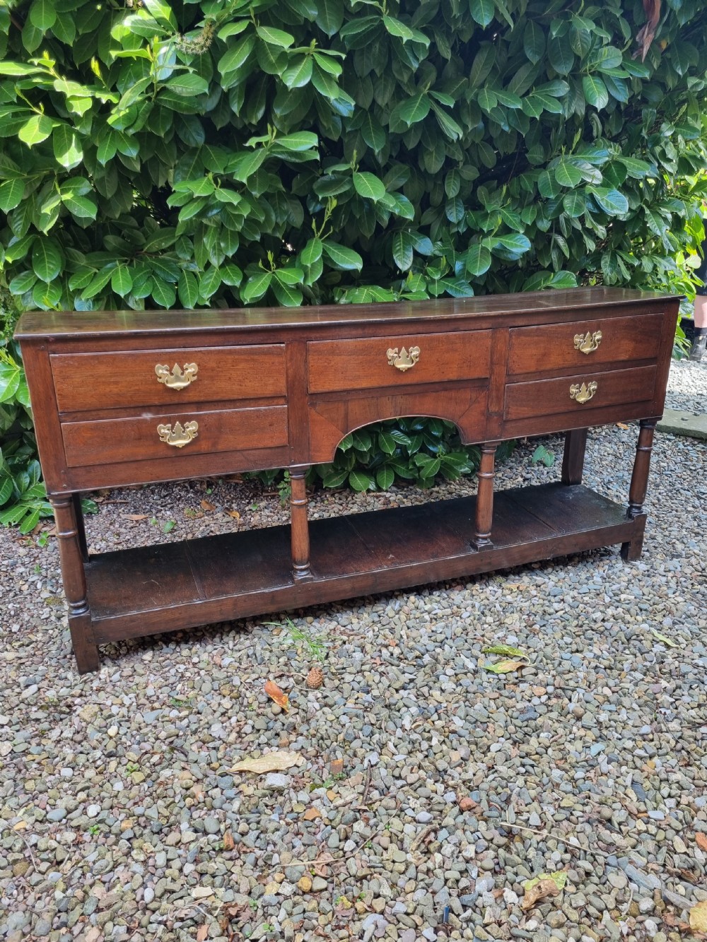 An eighteenth-century dark oak Welsh dresser base featuring five drawers with brass swan-neck handles, an arched central apron, turned supports, and a lower pot board shelf. View VIII
