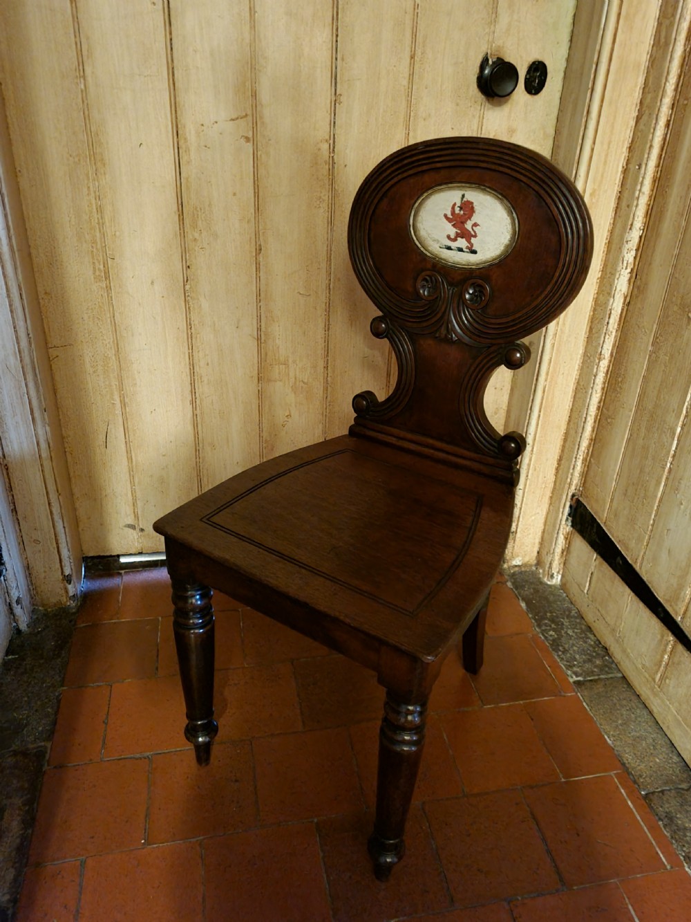 A Regency mahogany hall chair featuring an oval back panel with a central painted heraldic crest, a solid timber seat, and turned front legs.