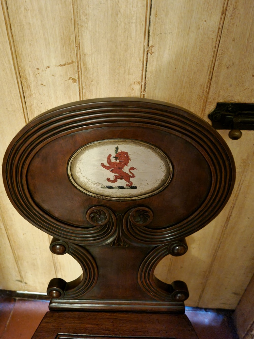 A Regency mahogany hall chair featuring an oval back panel with a central painted heraldic crest, a solid timber seat, and turned front legs. View II