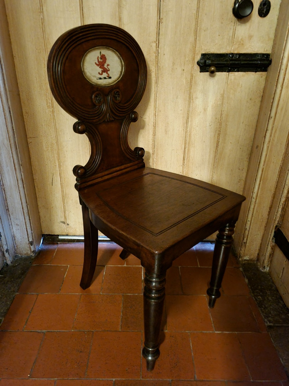 A Regency mahogany hall chair featuring an oval back panel with a central painted heraldic crest, a solid timber seat, and turned front legs. View III