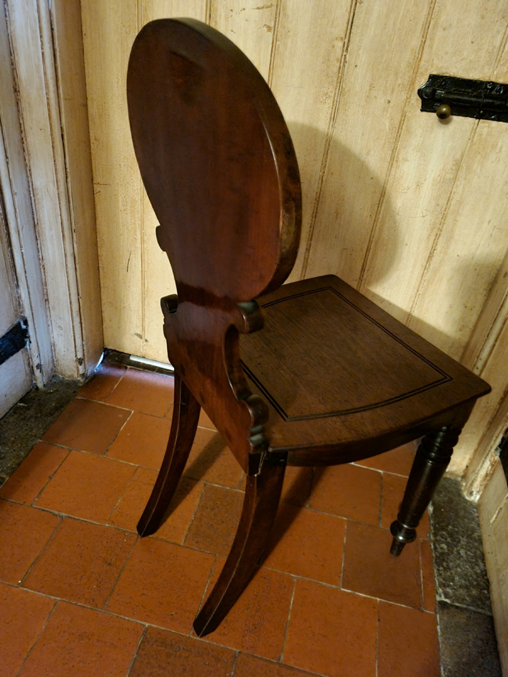 A Regency mahogany hall chair featuring an oval back panel with a central painted heraldic crest, a solid timber seat, and turned front legs. View V