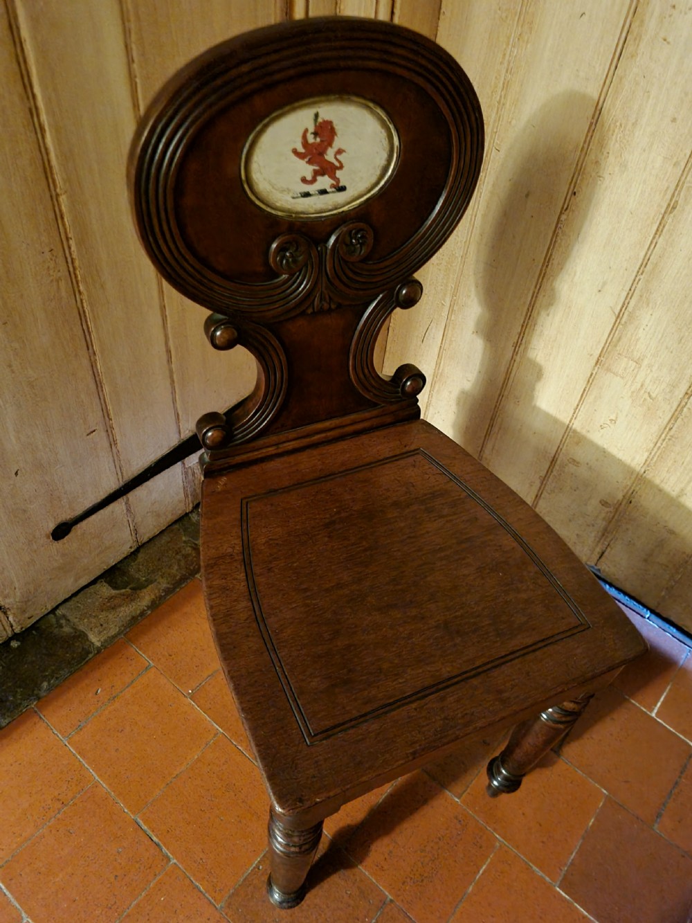 A Regency mahogany hall chair featuring an oval back panel with a central painted heraldic crest, a solid timber seat, and turned front legs. View VI