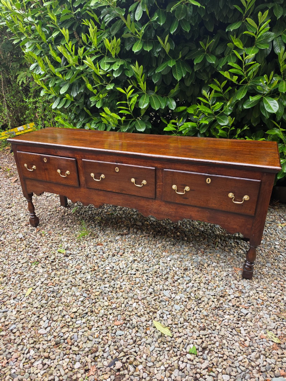 A mid-18th-century Georgian oak dresser base featuring three frieze drawers with brass swan-neck handles, a shaped apron, and turned front legs.
